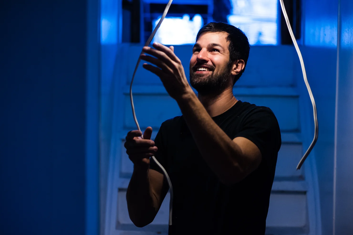 A smiling person is holding cables in a room with blue lighting. They appear engaged and happy, wearing a black shirt, against a dark background.