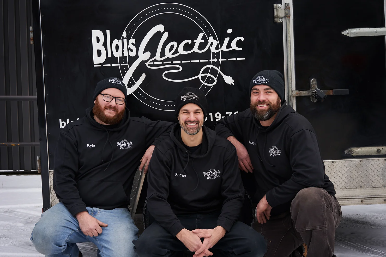 Three smiling people in matching black hoodies pose in front of a trailer with "Blais Electric" logo. They appear to be colleagues.