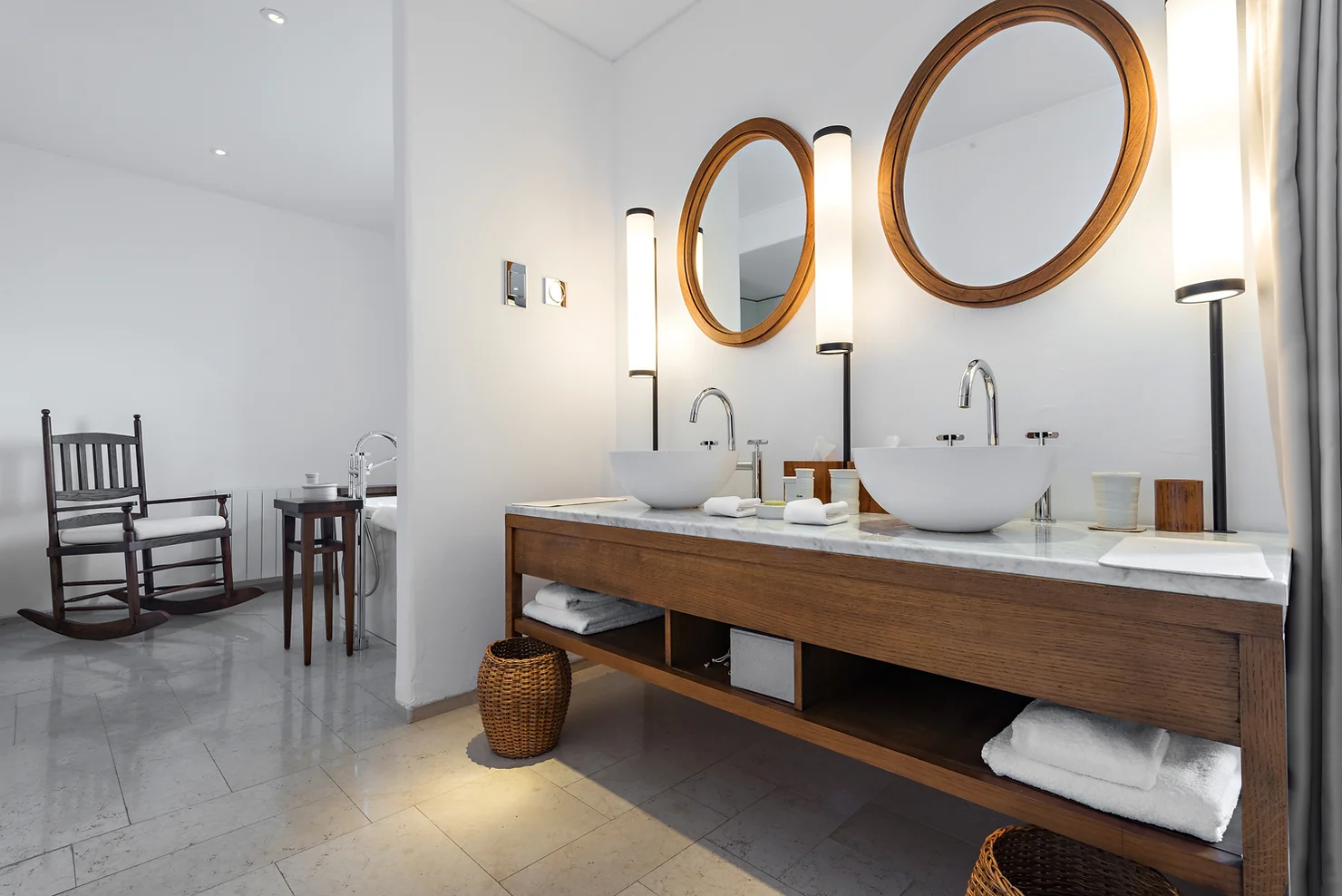 A modern bathroom features twin round mirrors, two vessel sinks, long pendant lights, and a wooden vanity with towels beneath. A rocking chair nearby.