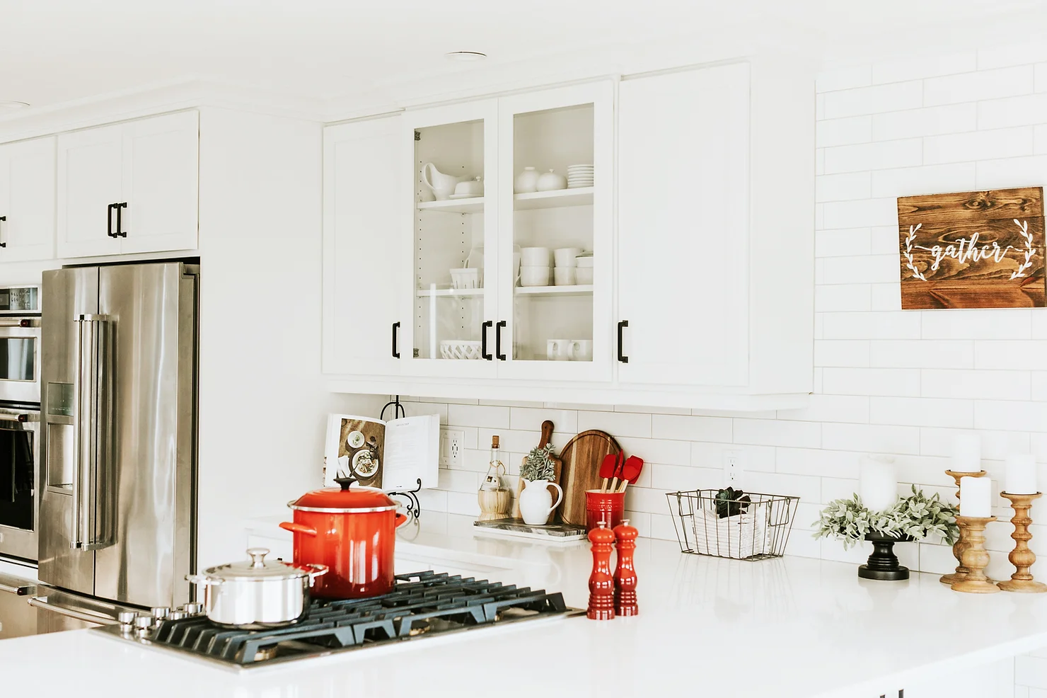 Modern kitchen interior with white cabinetry, stainless steel appliances, a gas stove, red pots, and decorative elements. A wooden sign reads "gather."
