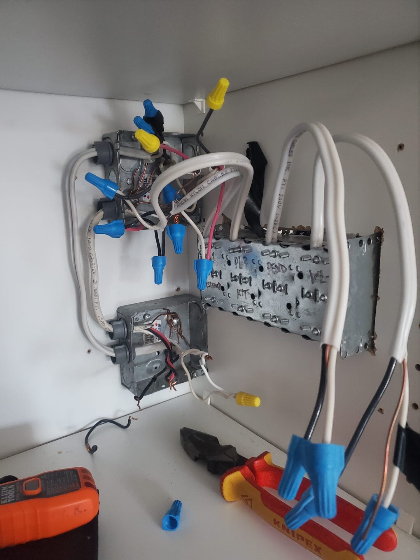 An electrical installation in progress with wires, wire nuts, junction boxes, and tools like screwdrivers and a voltage tester on a white surface.
