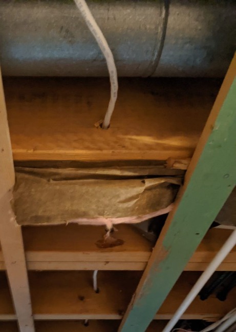 The image shows an interior space beneath a large duct with damaged and sagging insulation. Exposed wooden beams and pipes are also visible.