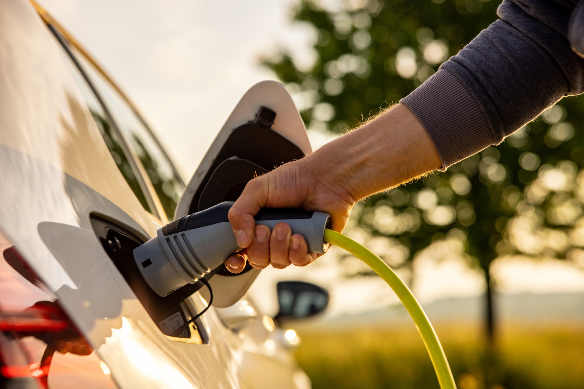 A person's hand is connecting an electric vehicle charger to a white car during sunrise or sunset, with a natural background and clear skies.