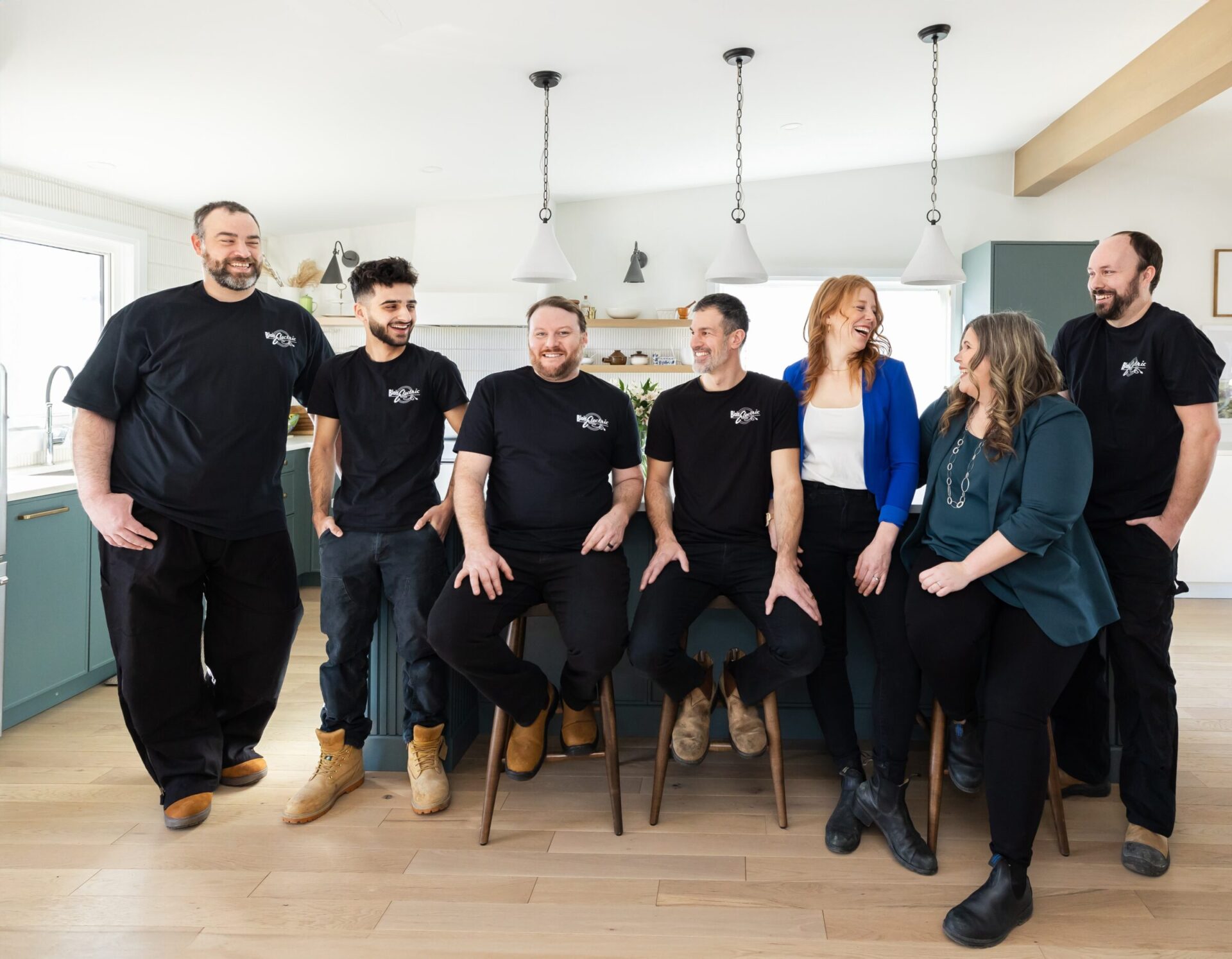 Seven people, some seated on stools, pose smiling in a bright, modern kitchen. Most wear matching black shirts with a logo, suggesting a unified team.