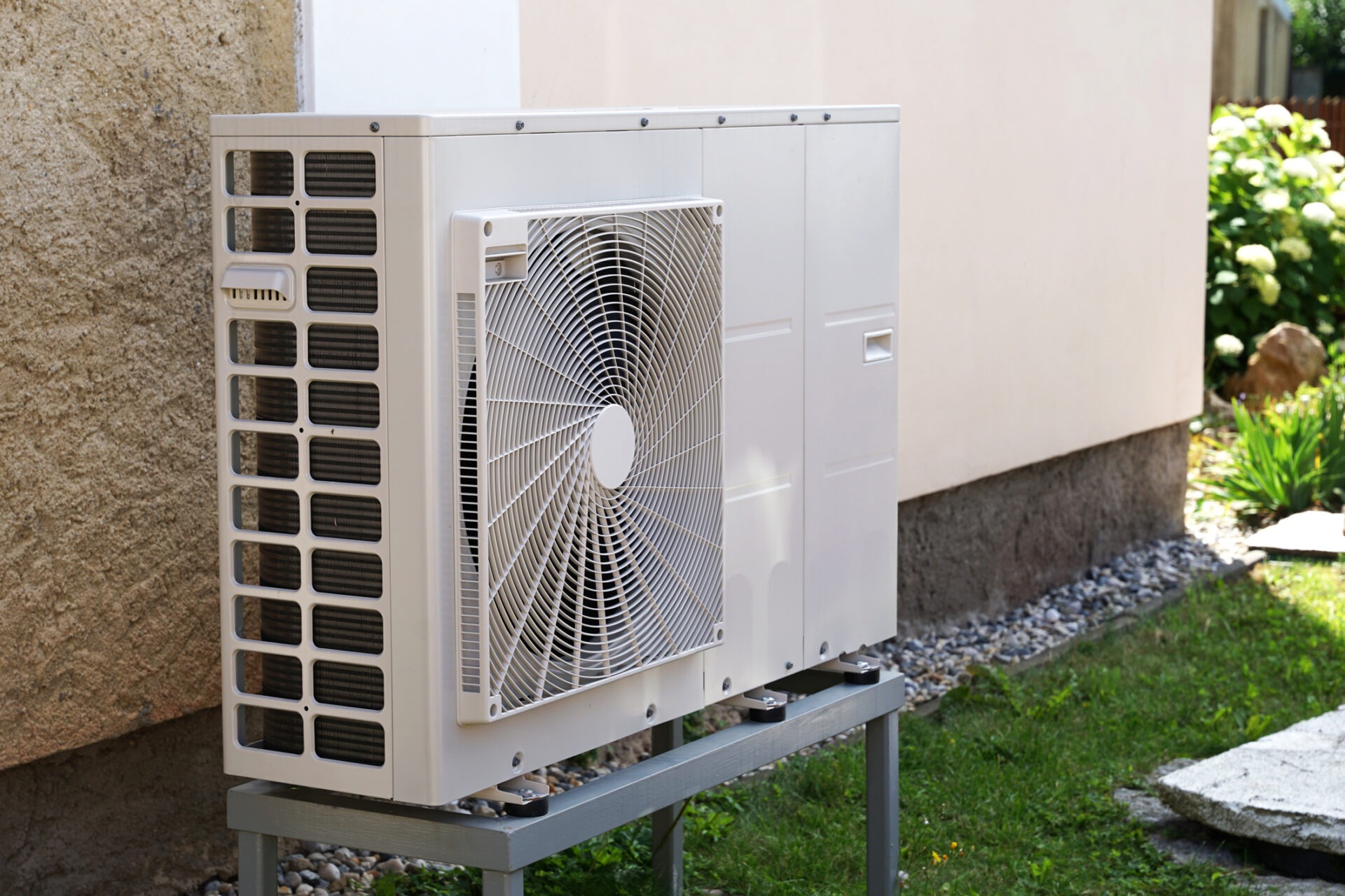 An outdoor heat pump unit is installed on a metal stand beside a house, with a well-manicured lawn and plants in the background.