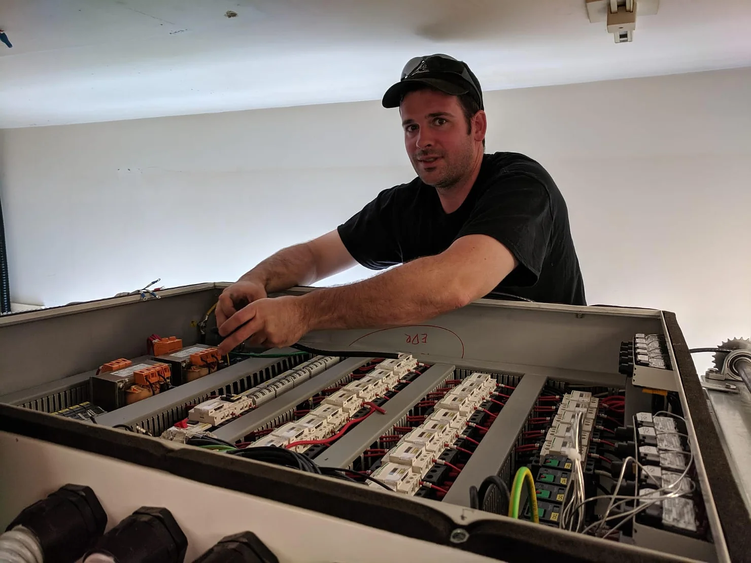 A person wearing a cap is working on an industrial electrical panel with various switches and wiring, looking towards the camera.