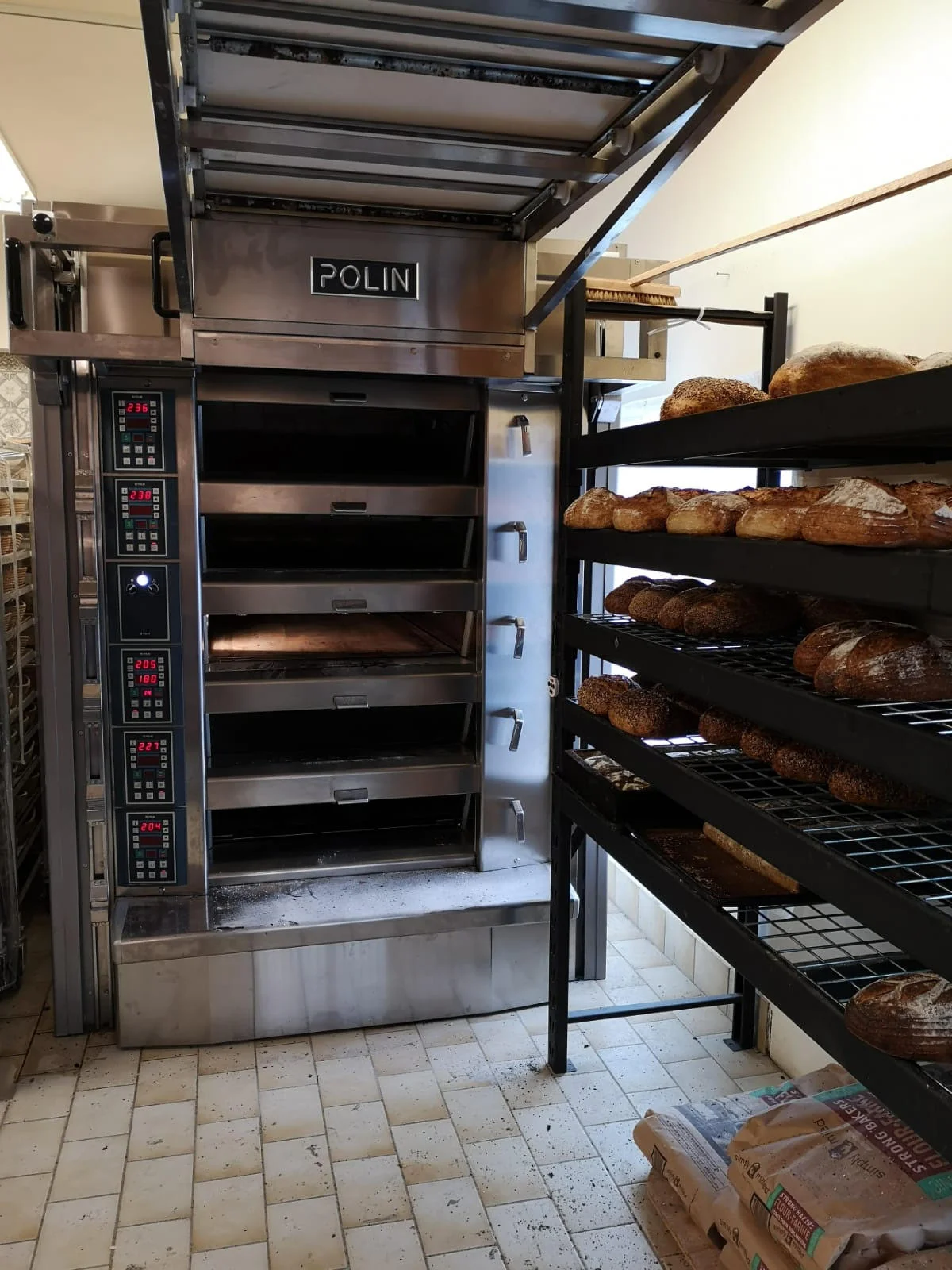 A professional bakery oven with multiple slots is open, displaying empty racks. Various baked breads are cooling off on a shelf beside it.