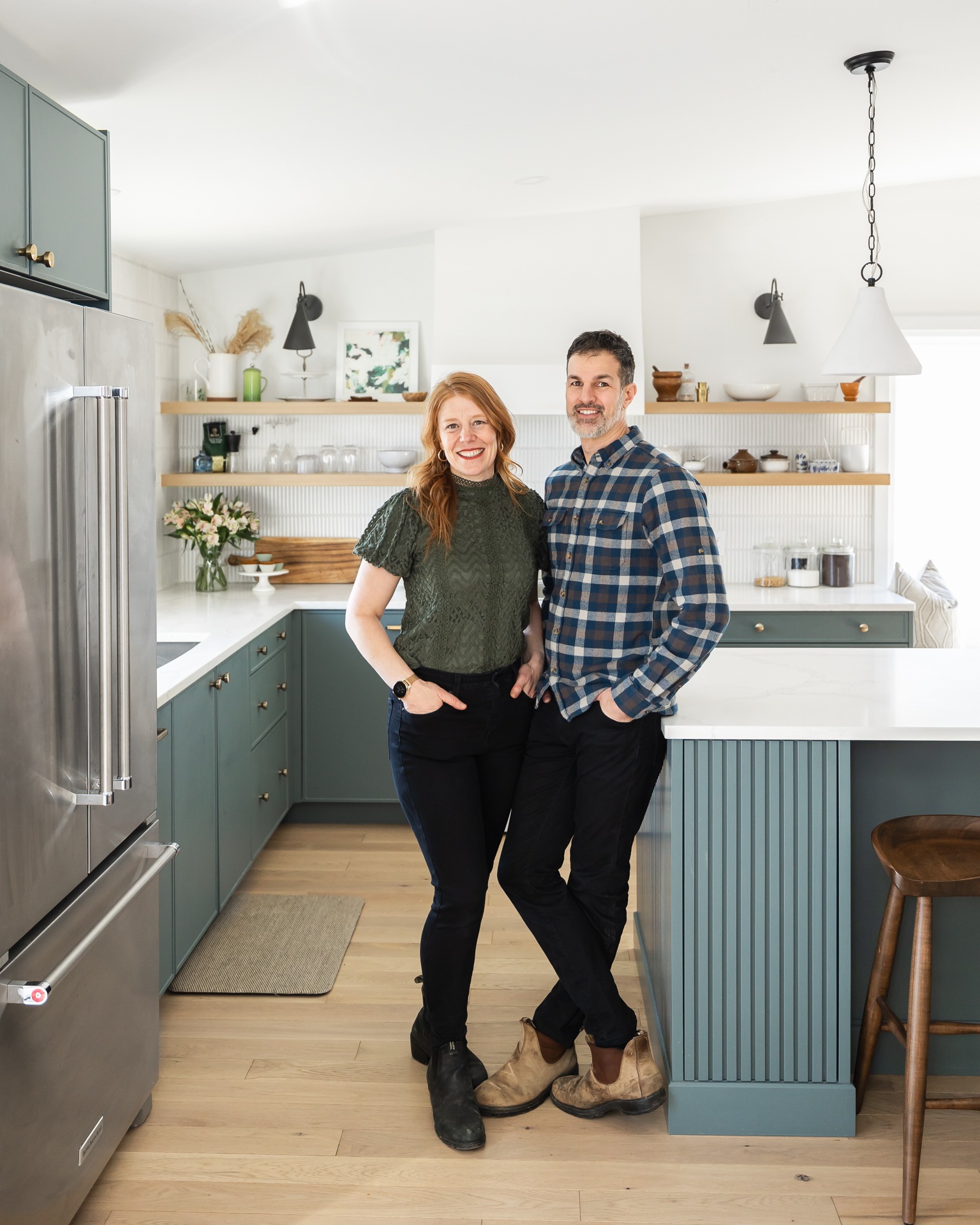 Two people are standing in a modern kitchen with teal cabinetry, smiling at the camera. There's an island, open shelves, and stainless steel appliances.