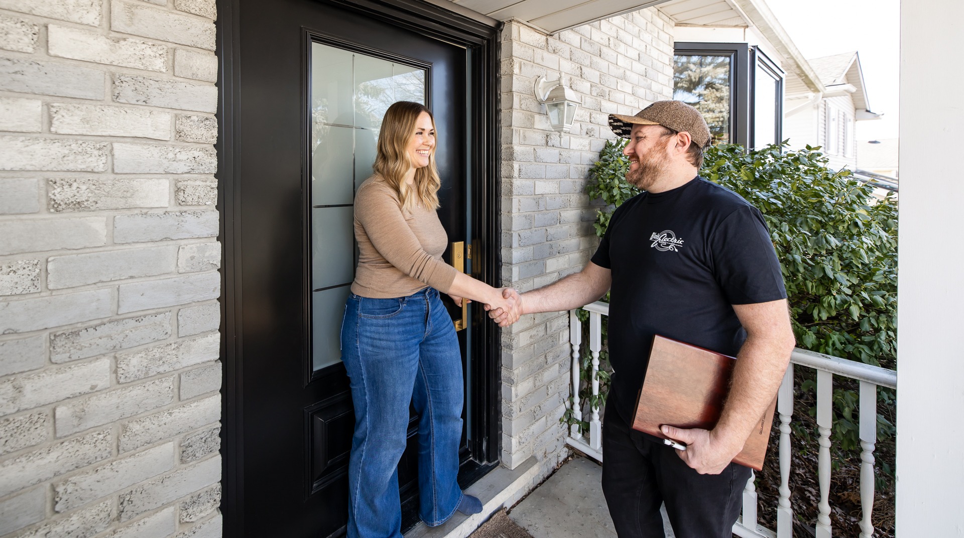 A person is greeting another at the front door; they are shaking hands, smiling, with bright daylight illuminating a residential entrance.
