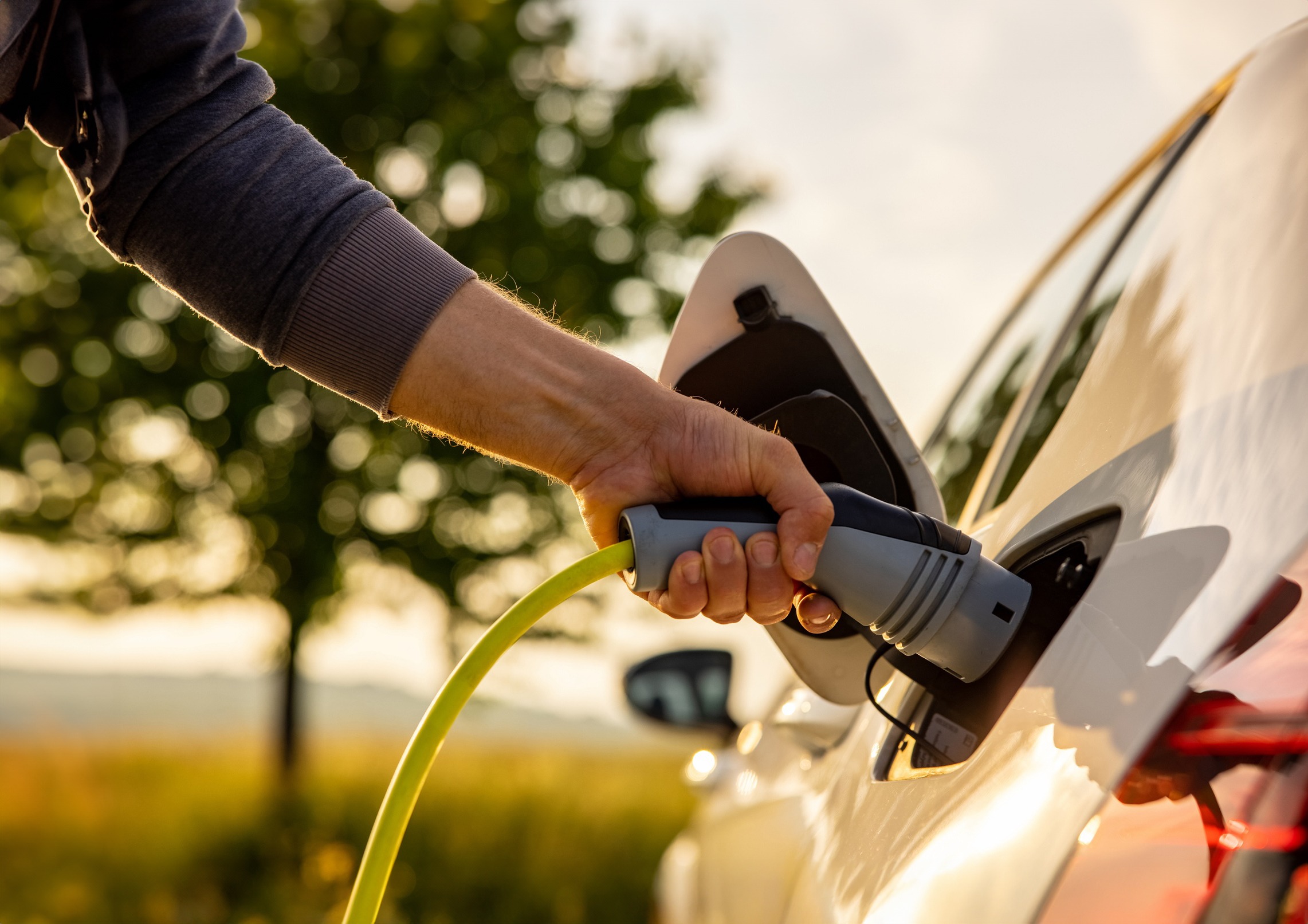 A person is plugging an electric vehicle charging connector into a white car, with a backdrop of a sunlit sky and trees.