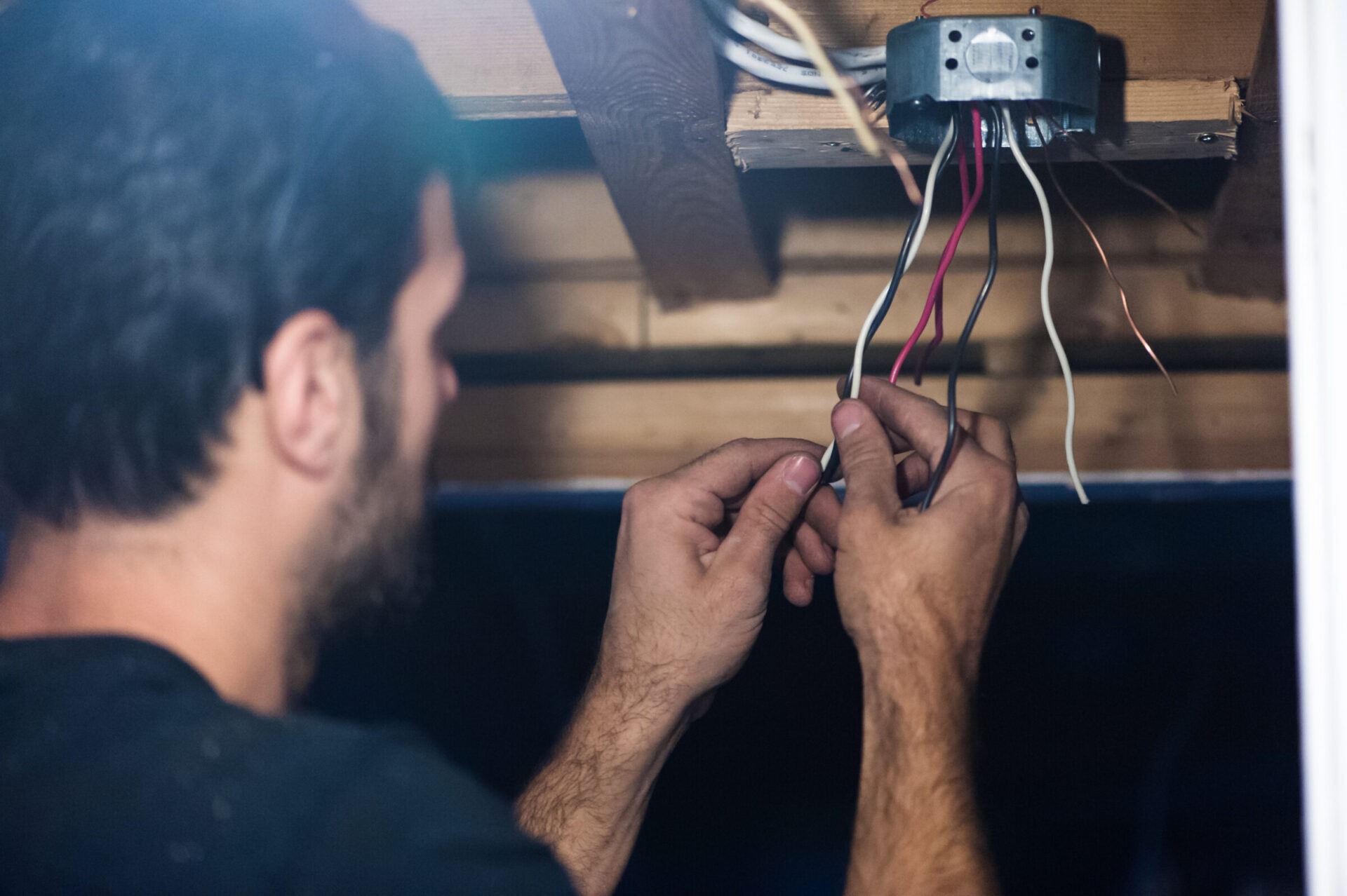 A person is installing or repairing electrical wiring, focused on connecting wires in a junction box in a dimly lit, indoor setting.