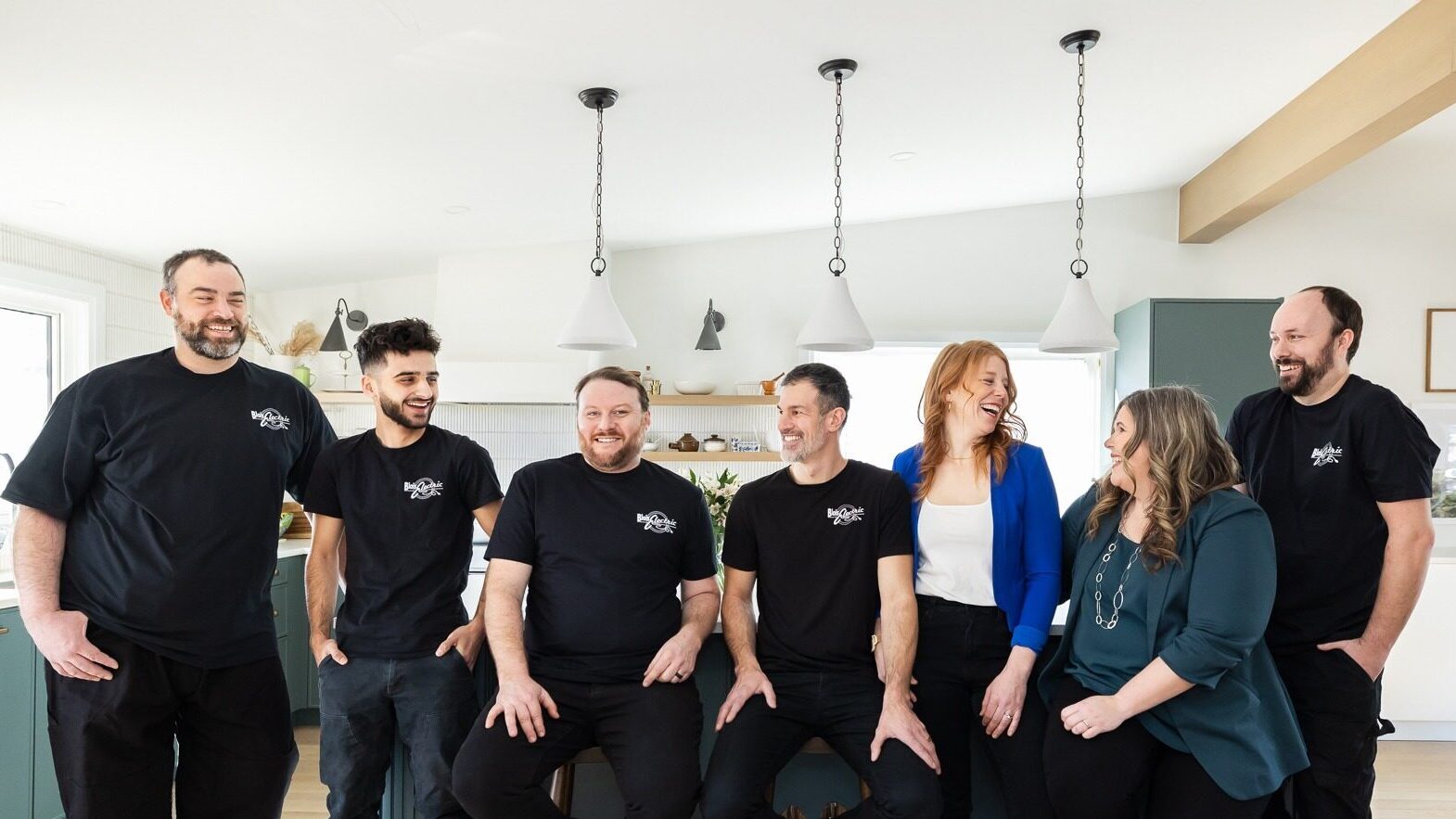 Seven adults stand and sit in a modern kitchen, smiling and wearing black t-shirts with a white logo, looking relaxed and happy.