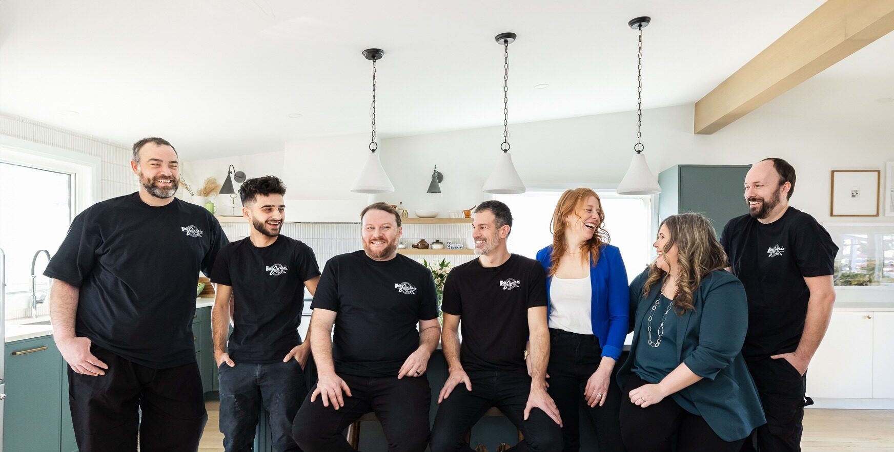 Eight people, wearing matching black T-shirts with a logo, are smiling and posing together in a bright, modern kitchen with pendant lights.