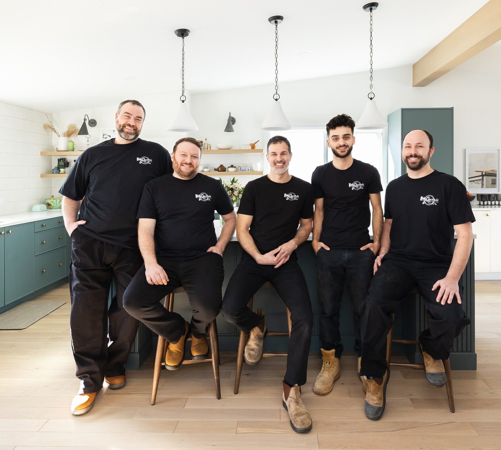 Five people wearing black shirts with matching logos pose smiling in a bright kitchen with teal cabinets, pendant lights, and a wooden counter.