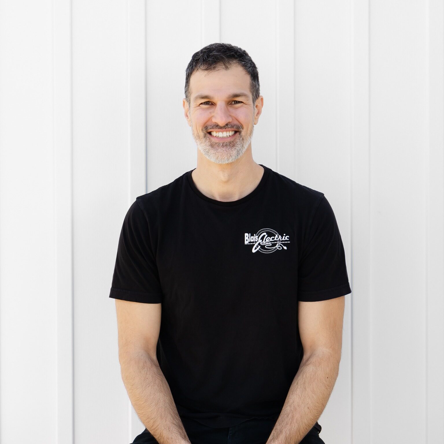 A person with a beard smiling, wearing a black t-shirt, standing against a white vertical plank background, looking relaxed and happy.
