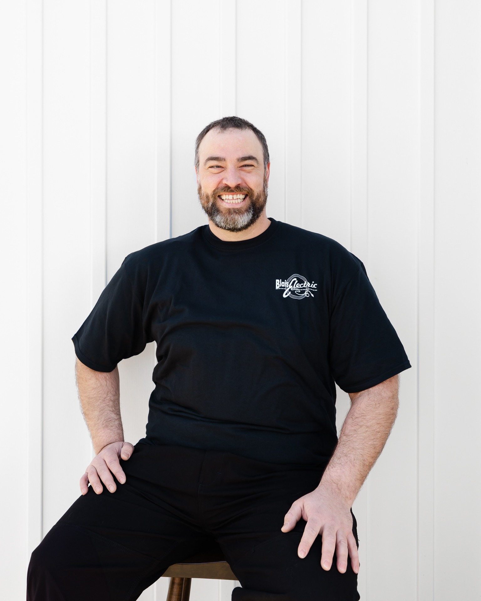 A person with a beard smiles, sitting relaxed on a stool, wearing a black T-shirt with a logo, against a white vertical siding background.