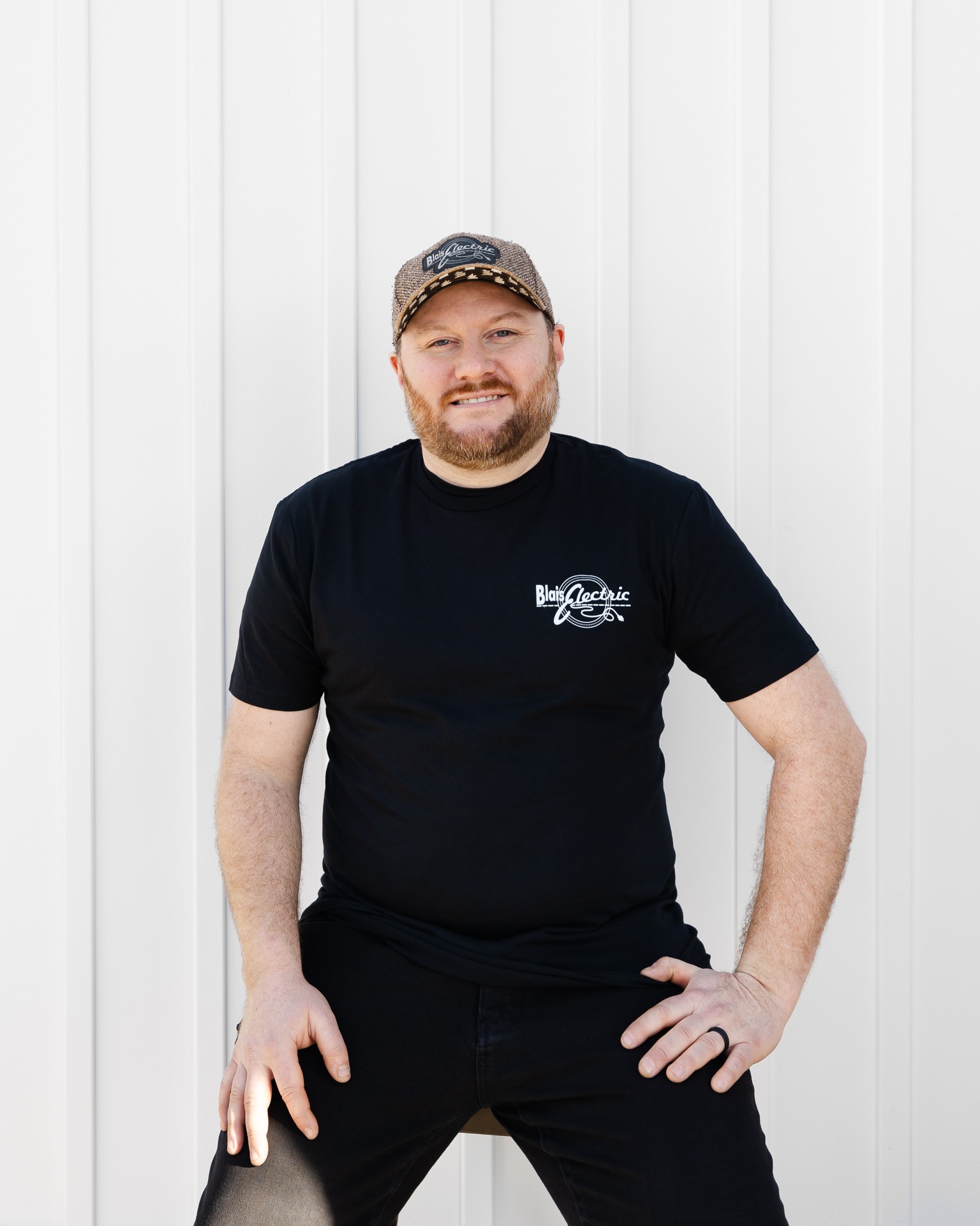 A person with a beard and cap stands confidently against a white corrugated wall, wearing a black t-shirt and pants, smiling slightly.