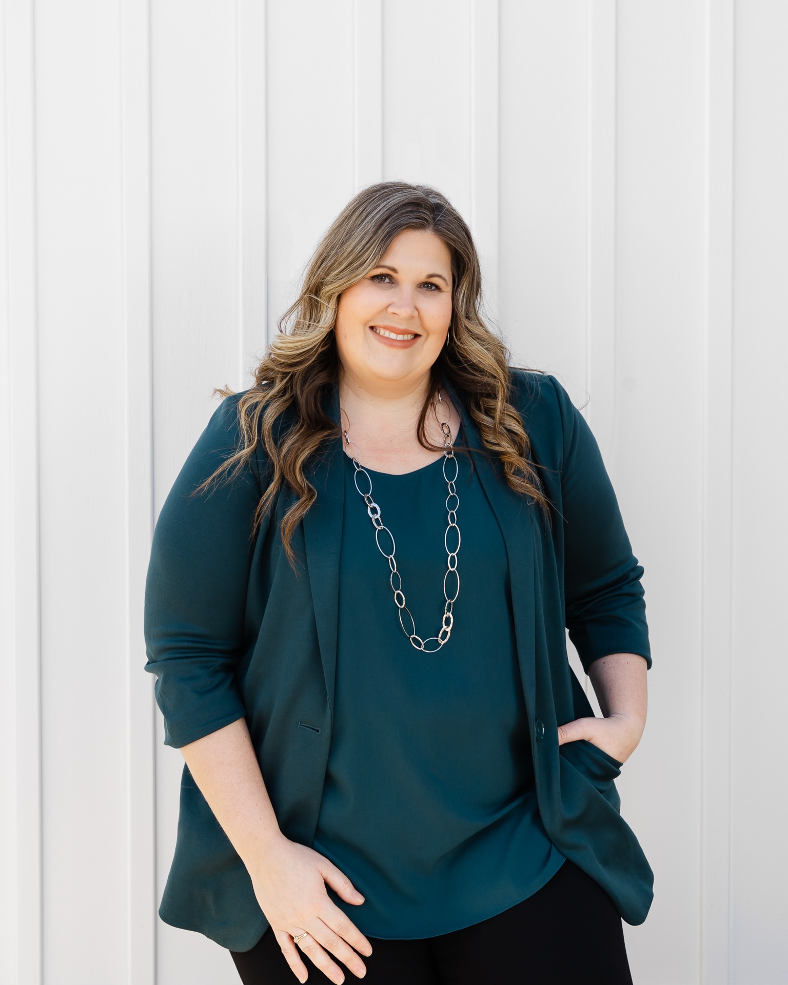 A smiling person stands confidently against a white vertical siding background, wearing a teal top, black pants, and a silver necklace.