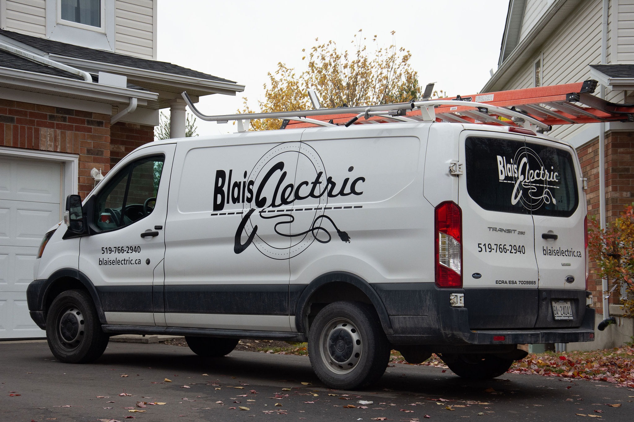 A white commercial van with "Blais Electric" decals parked in a driveway, with a ladder on the roof and autumn leaves on the ground.