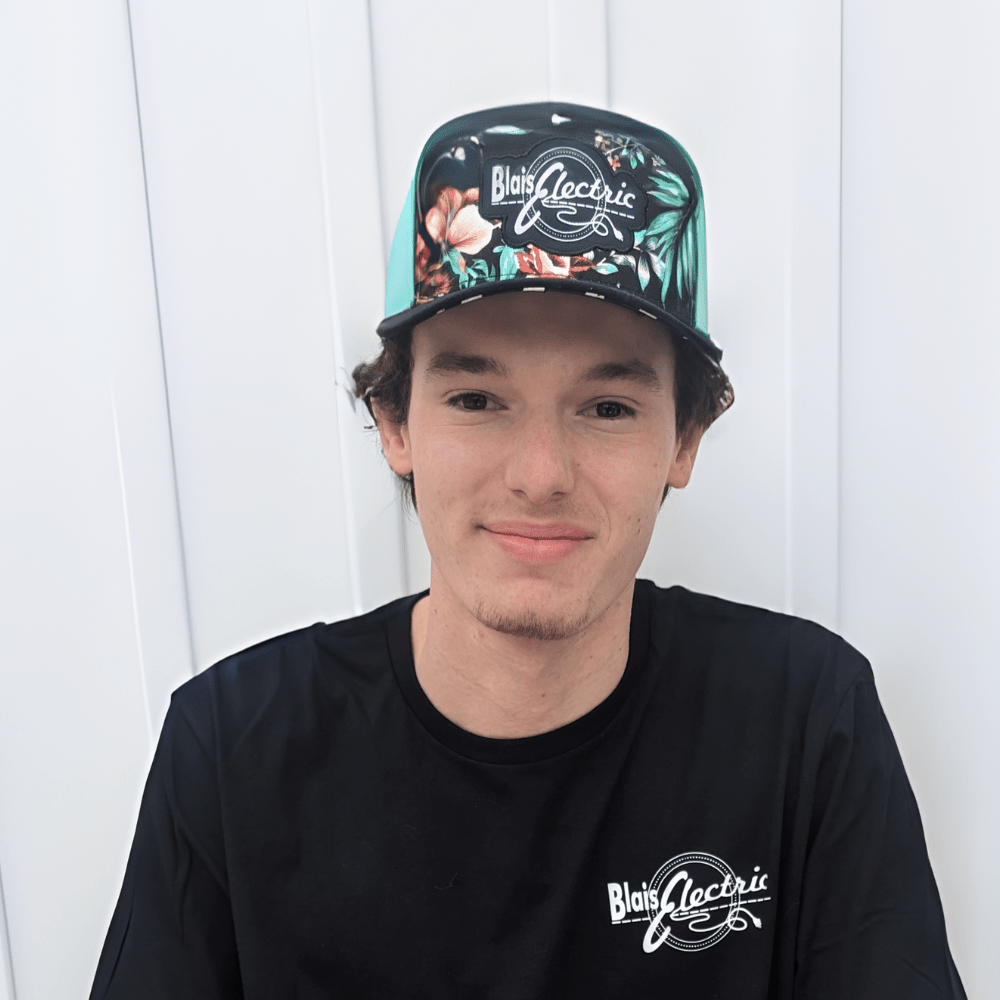 A person is smiling slightly, wearing a floral print cap and black t-shirt with graphics. Behind them is a white vertical panel background.