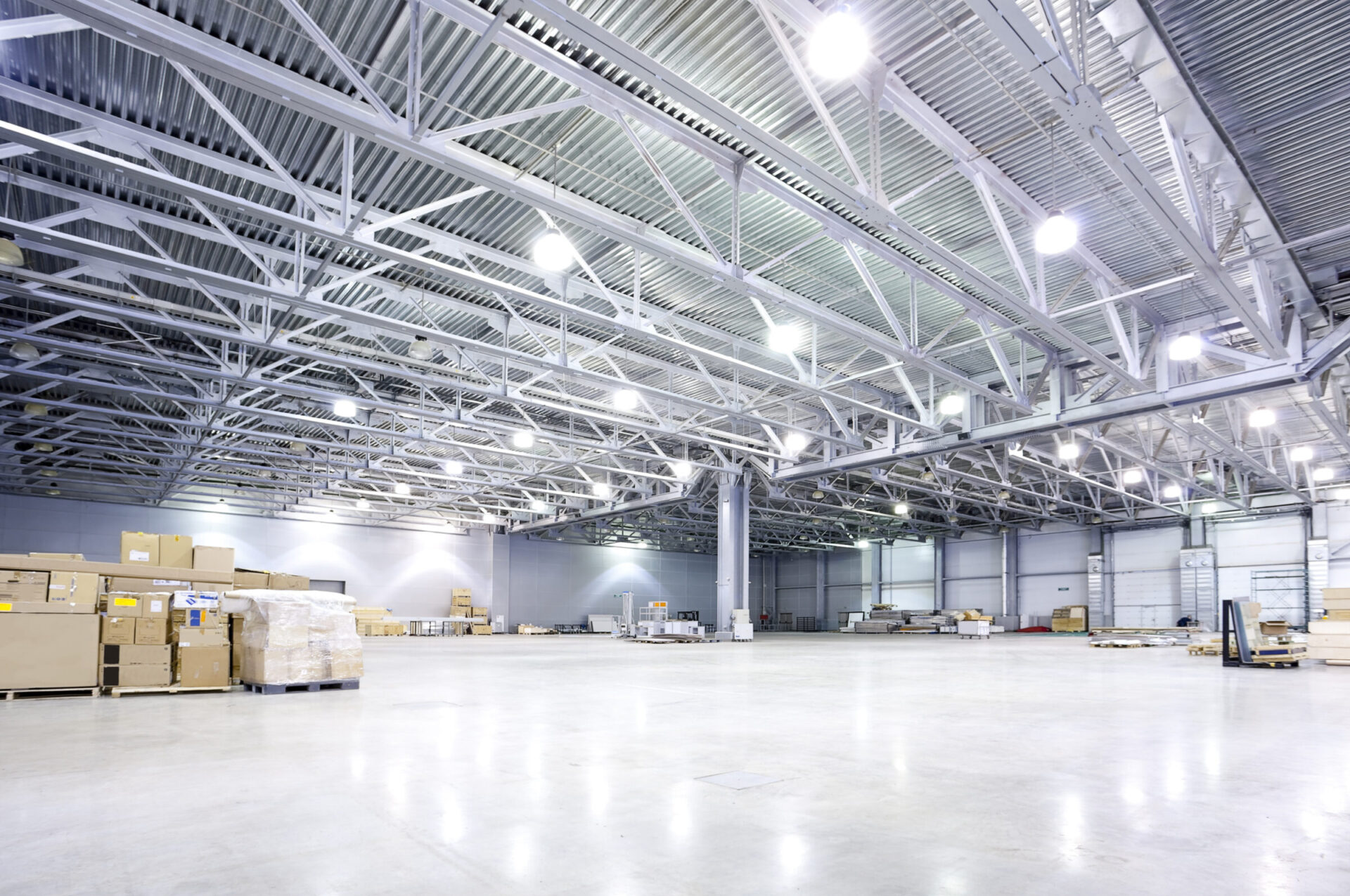 The image shows a spacious and brightly lit empty warehouse with high ceilings, metal trusses, and scattered boxes on palettes.