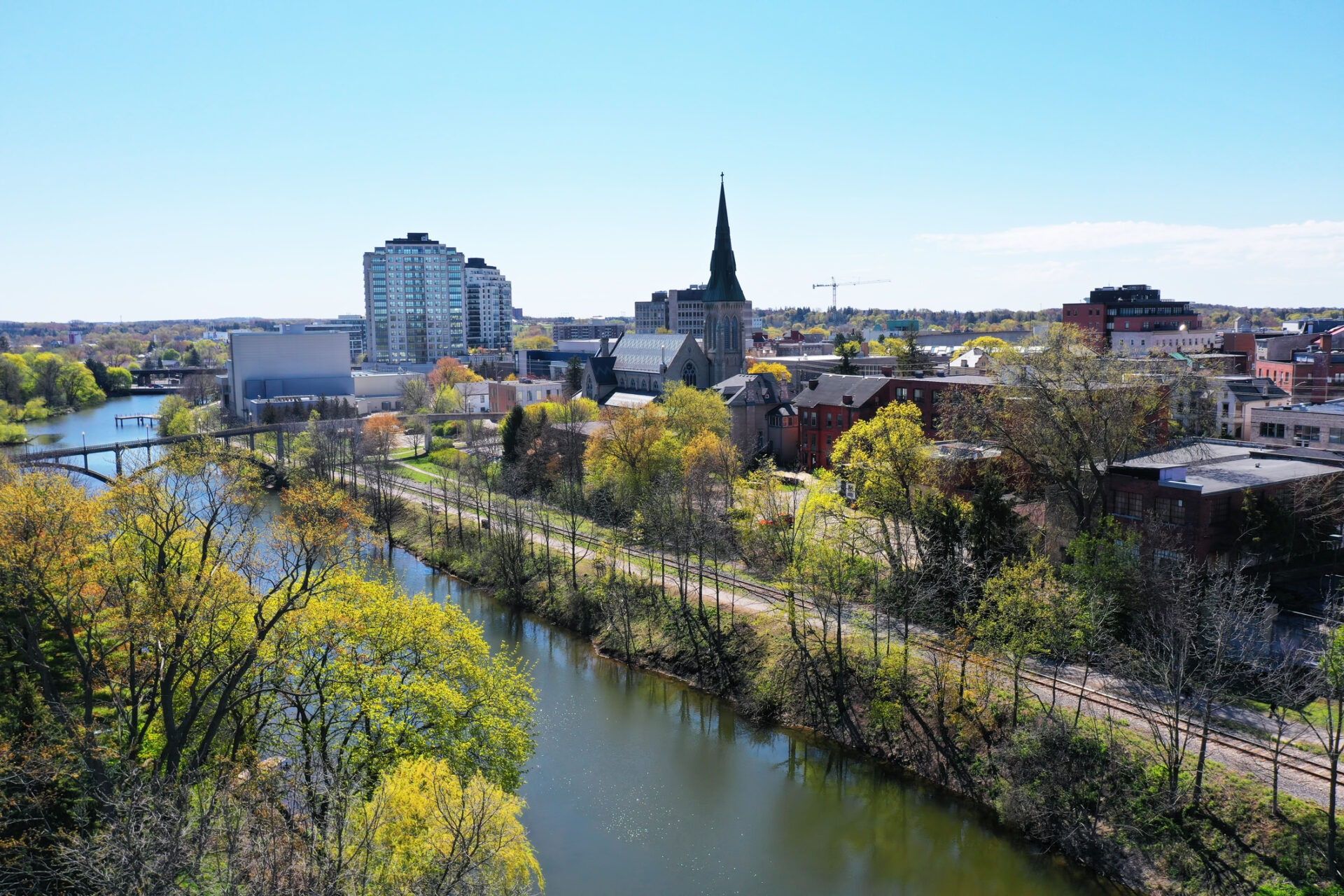 An aerial view of an urban landscape with a river, green foliage, a church spire, modern buildings, and clear blue skies.
