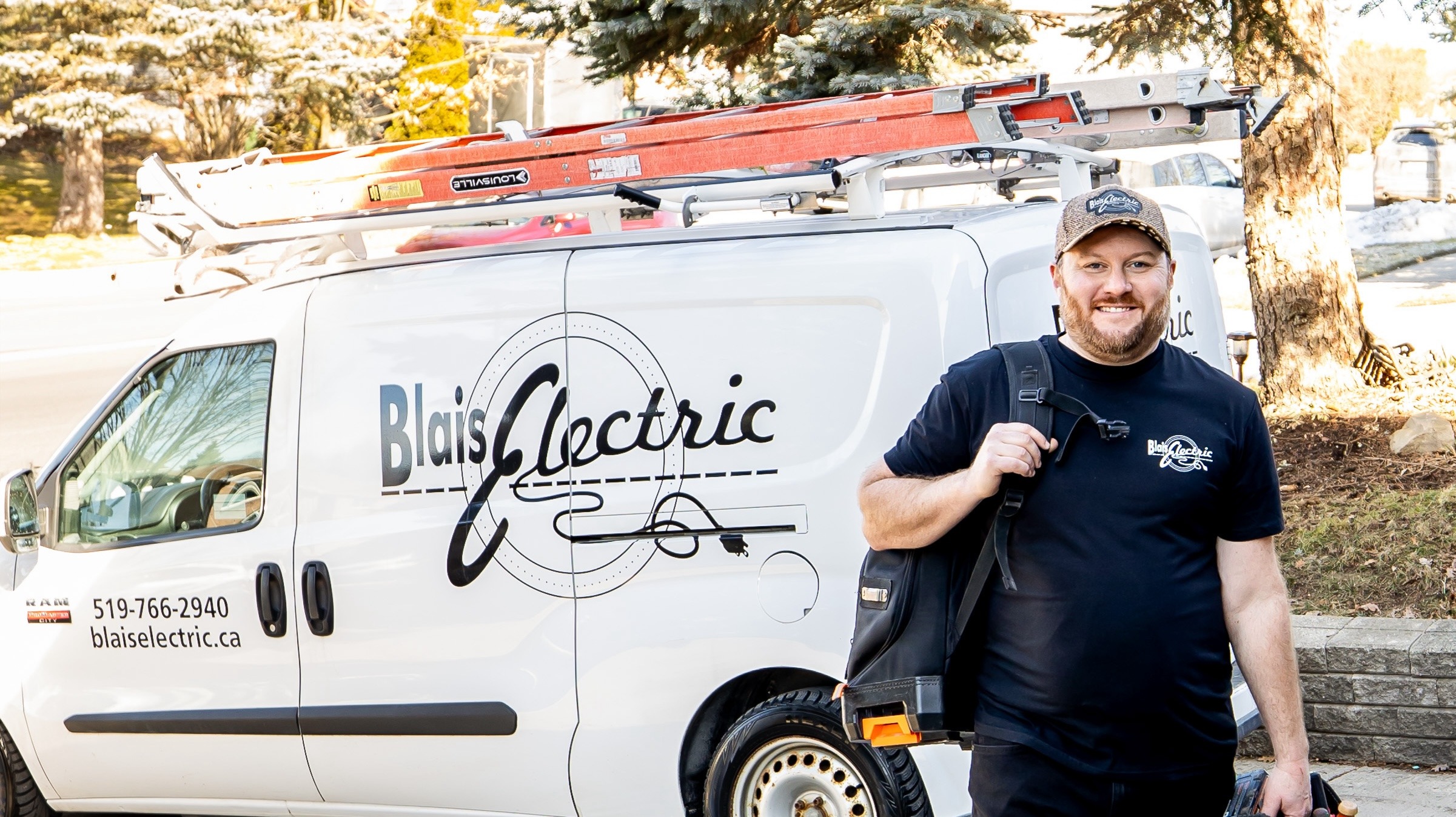 A smiling person stands beside a 'Blais Electric' van equipped with ladders, outdoors on a sunny day with trees and snow patches visible.