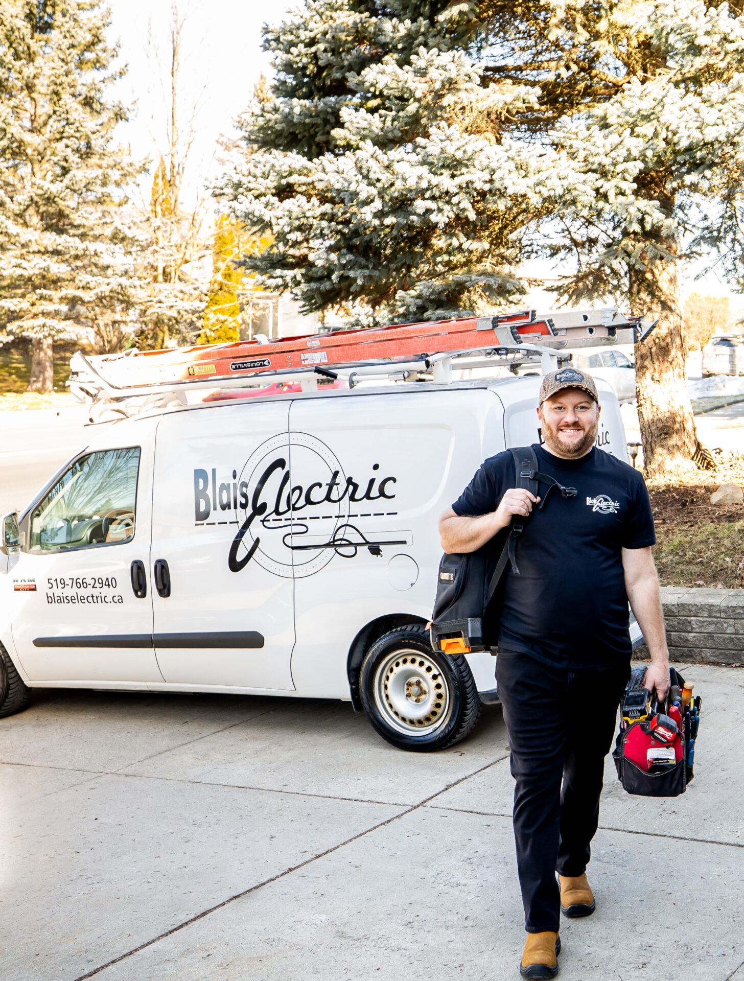 A smiling person stands in front of a white van labeled "Blais Electric," holding a tool bag, with a ladder on the van, and trees in the background.