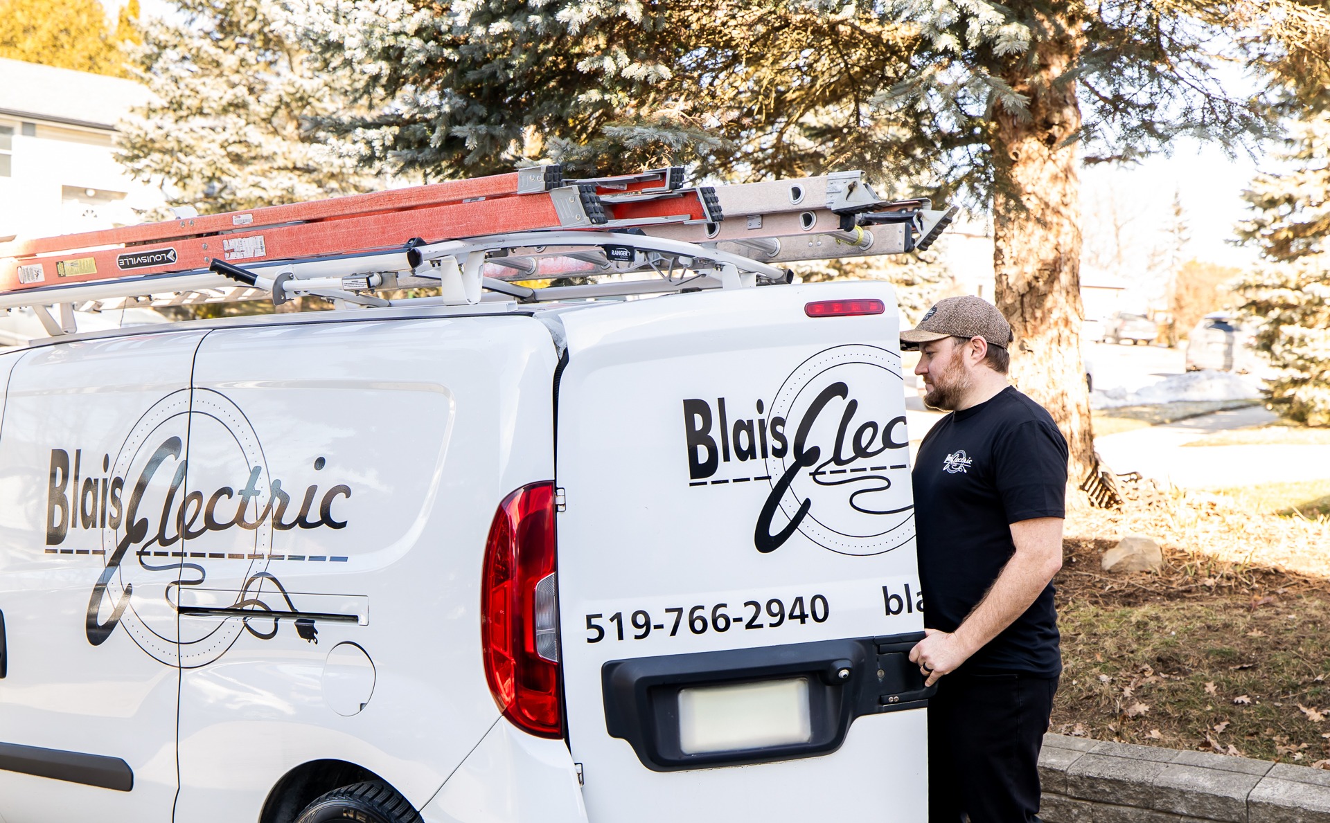 A person stands beside a white van with "Blais Electric" printed on it, equipped with ladders on top, in a residential area during daylight.