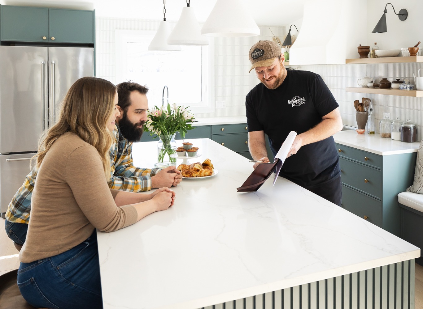 Three people are gathered in a bright, modern kitchen. One person reads a menu while two others sit at a counter with pastries and coffee.
