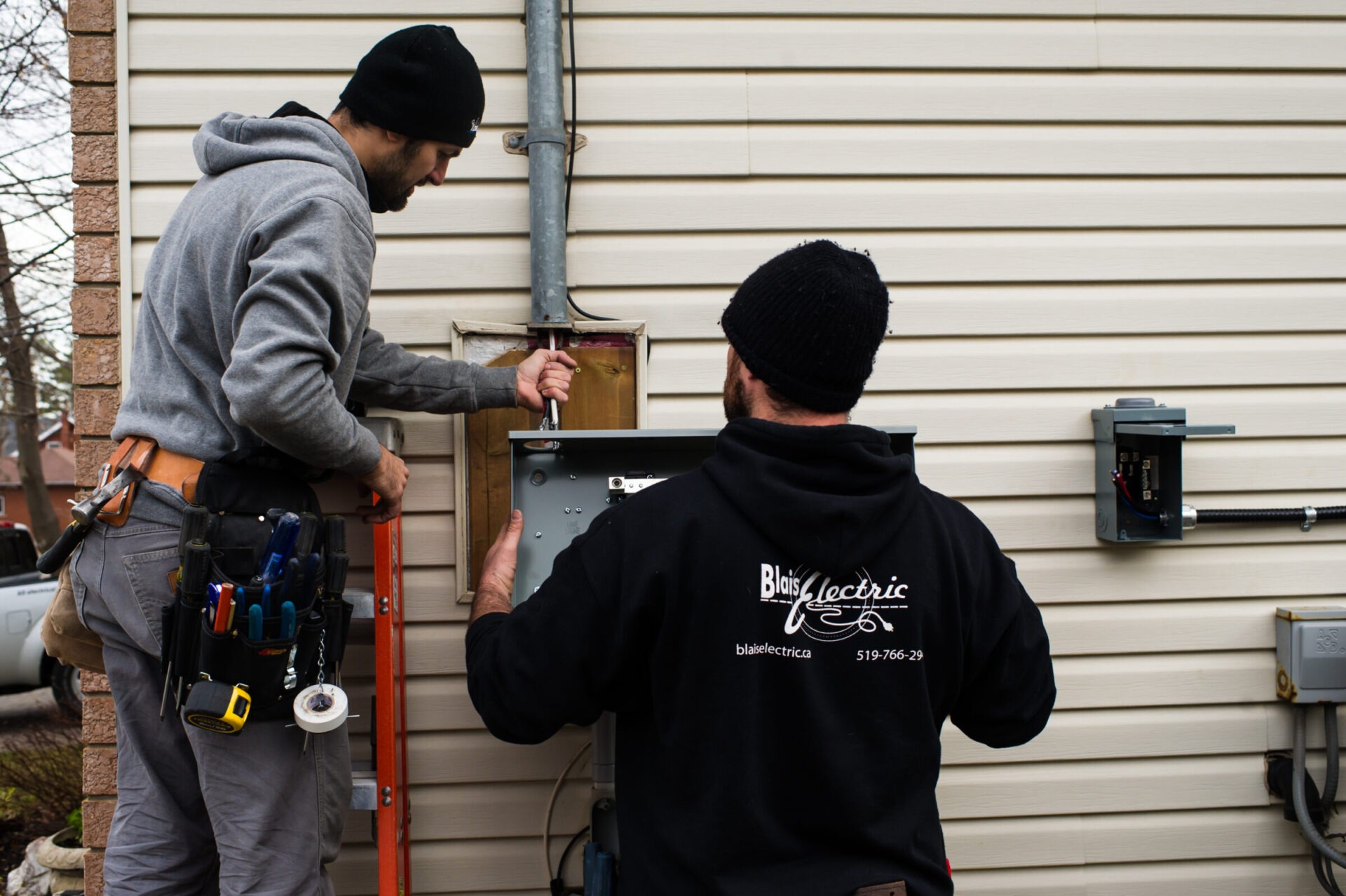 Two people are installing an electrical panel outdoors, one on a ladder. They are focused on their task, wearing casual clothes and utility belts.