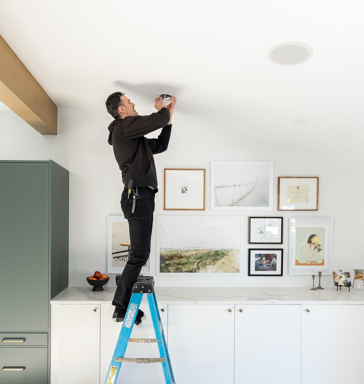 A person is standing on a ladder, installing a smoke detector on the ceiling of a kitchen adorned with framed artwork on the walls.