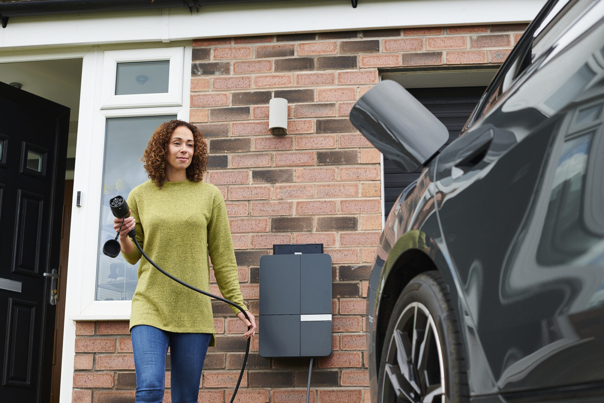 A person stands next to an electric vehicle, holding a charging plug near a home charging station outside a brick house with a white door.
