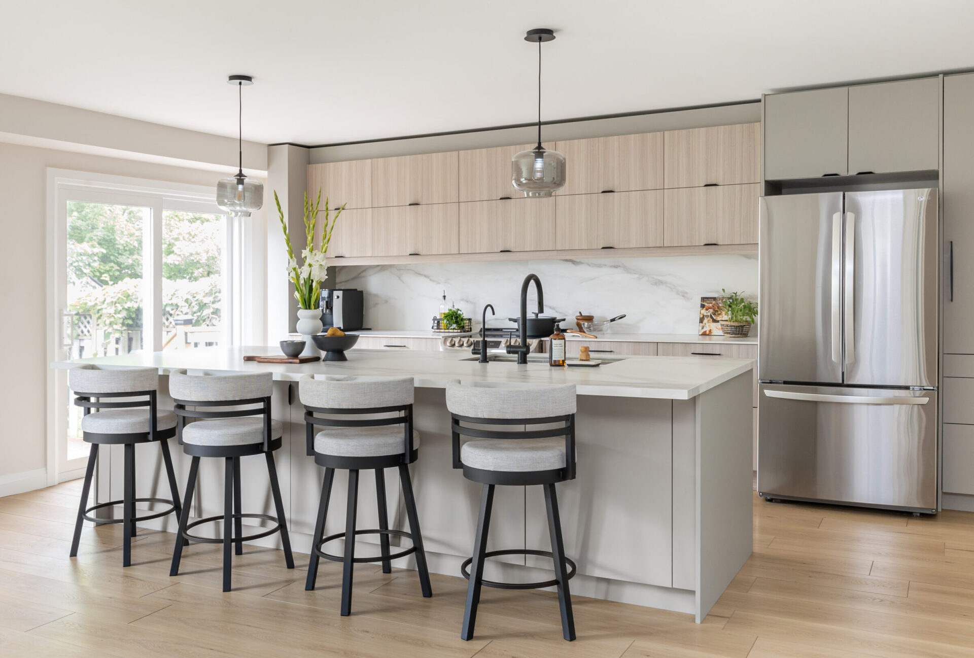 A modern kitchen interior with light wood cabinetry, stainless steel appliances, marble backsplash, and a white island with gray stools.