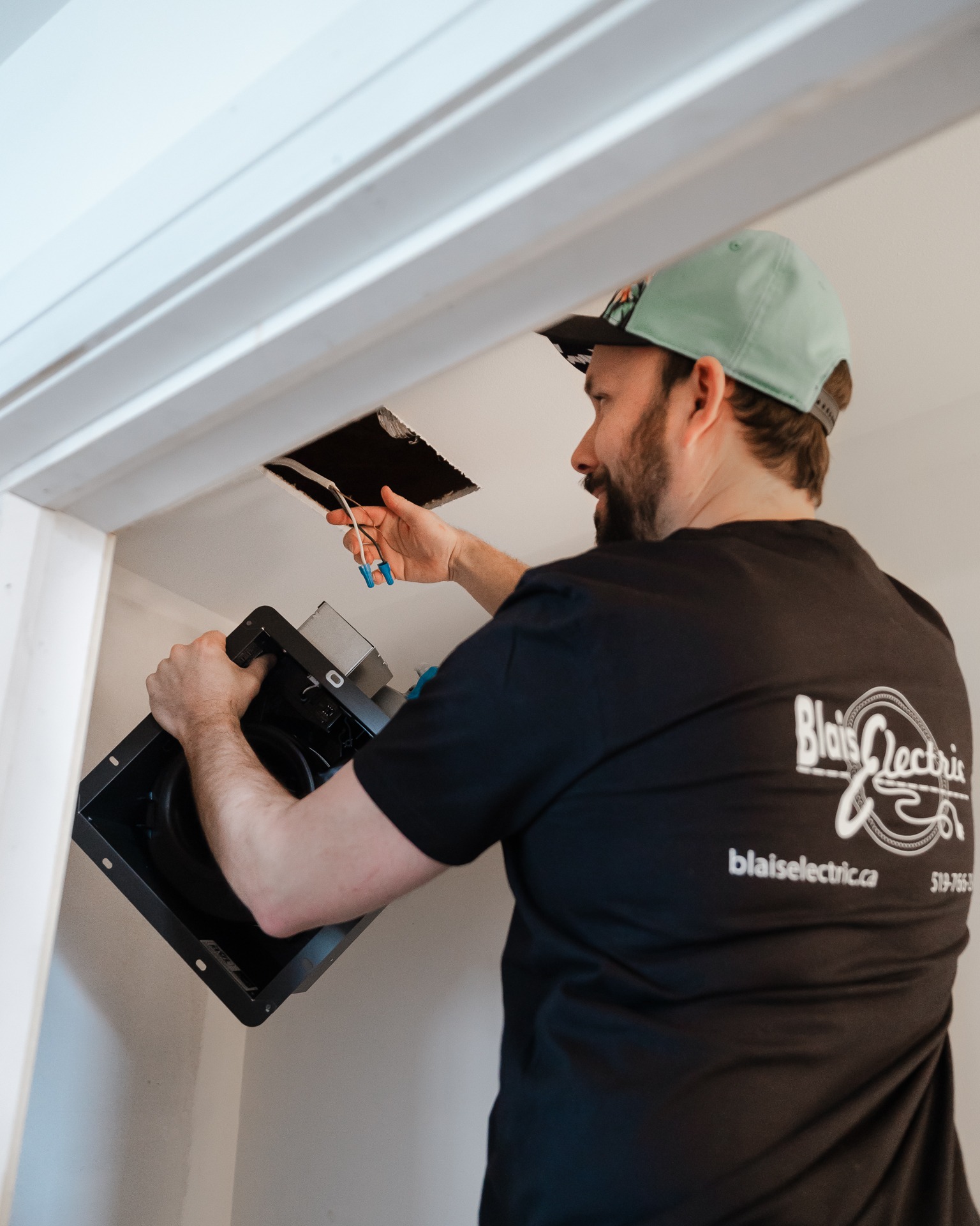 A person in a black shirt and baseball cap is installing a ceiling speaker, handling wires in a white interior setting.