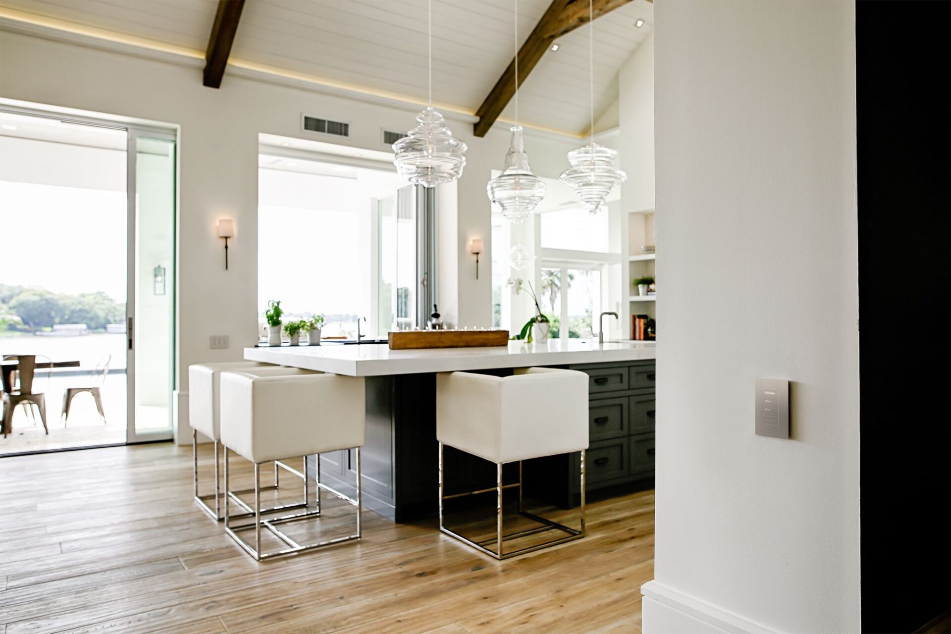 Modern kitchen interior with white bar stools at a black countertop island, three glass pendant lights, wooden floors, and a water view outdoors.