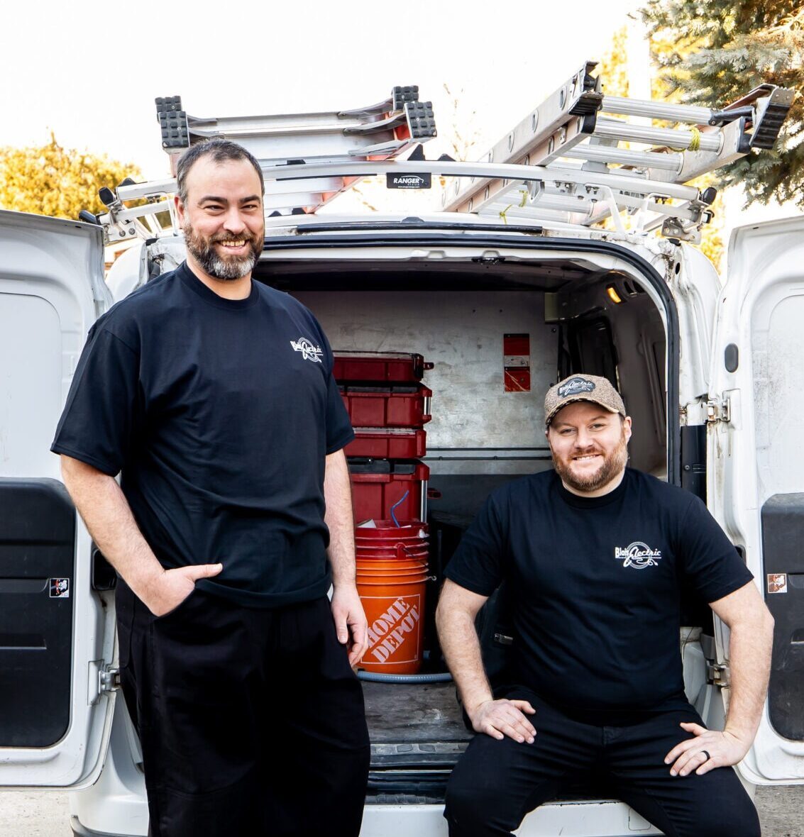 Two smiling persons in black shirts with company logos stand beside an open van filled with work equipment and red containers on a sunny day.