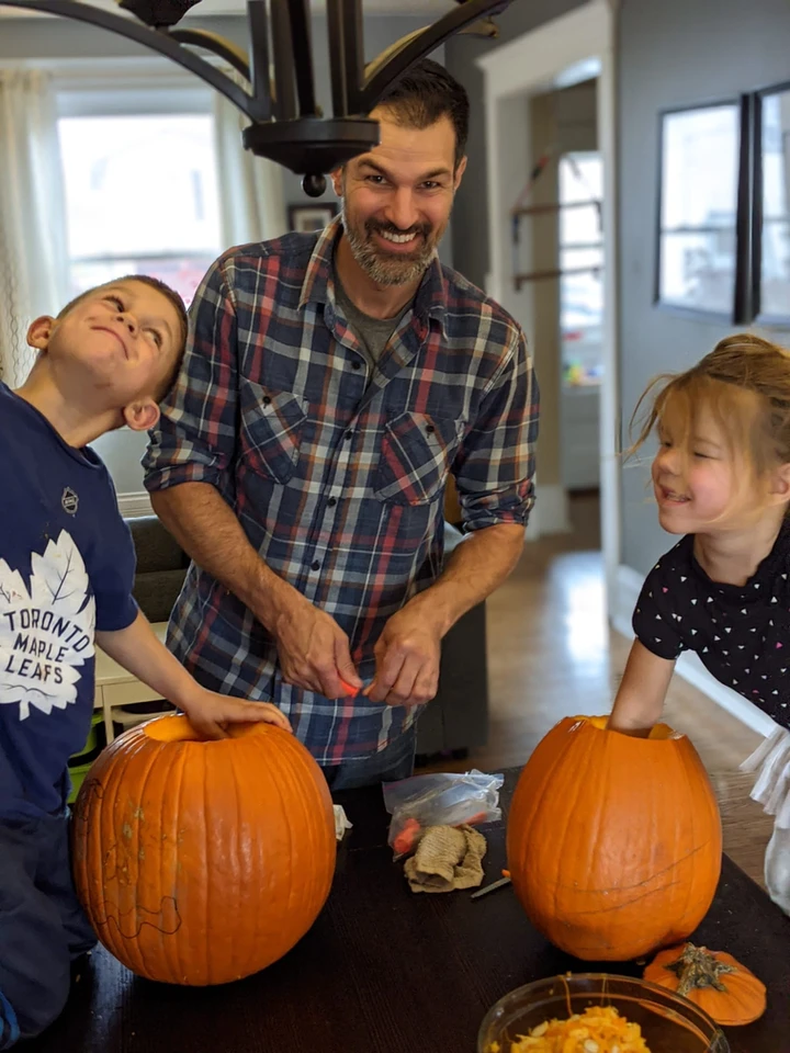A person and two children happily carve pumpkins at a table. The atmosphere is joyous, likely a family engaging in a Halloween tradition.