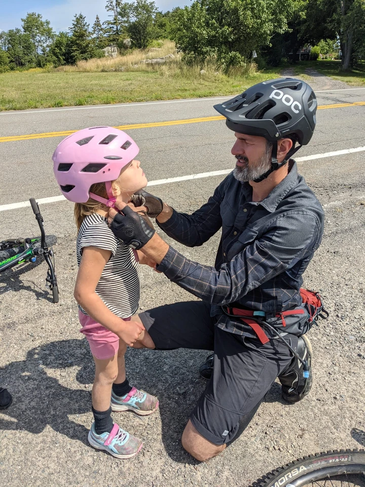 A person is helping a child with a pink helmet beside a bicycle on a sunny day, adjusting equipment, likely preparing for a bike ride.
