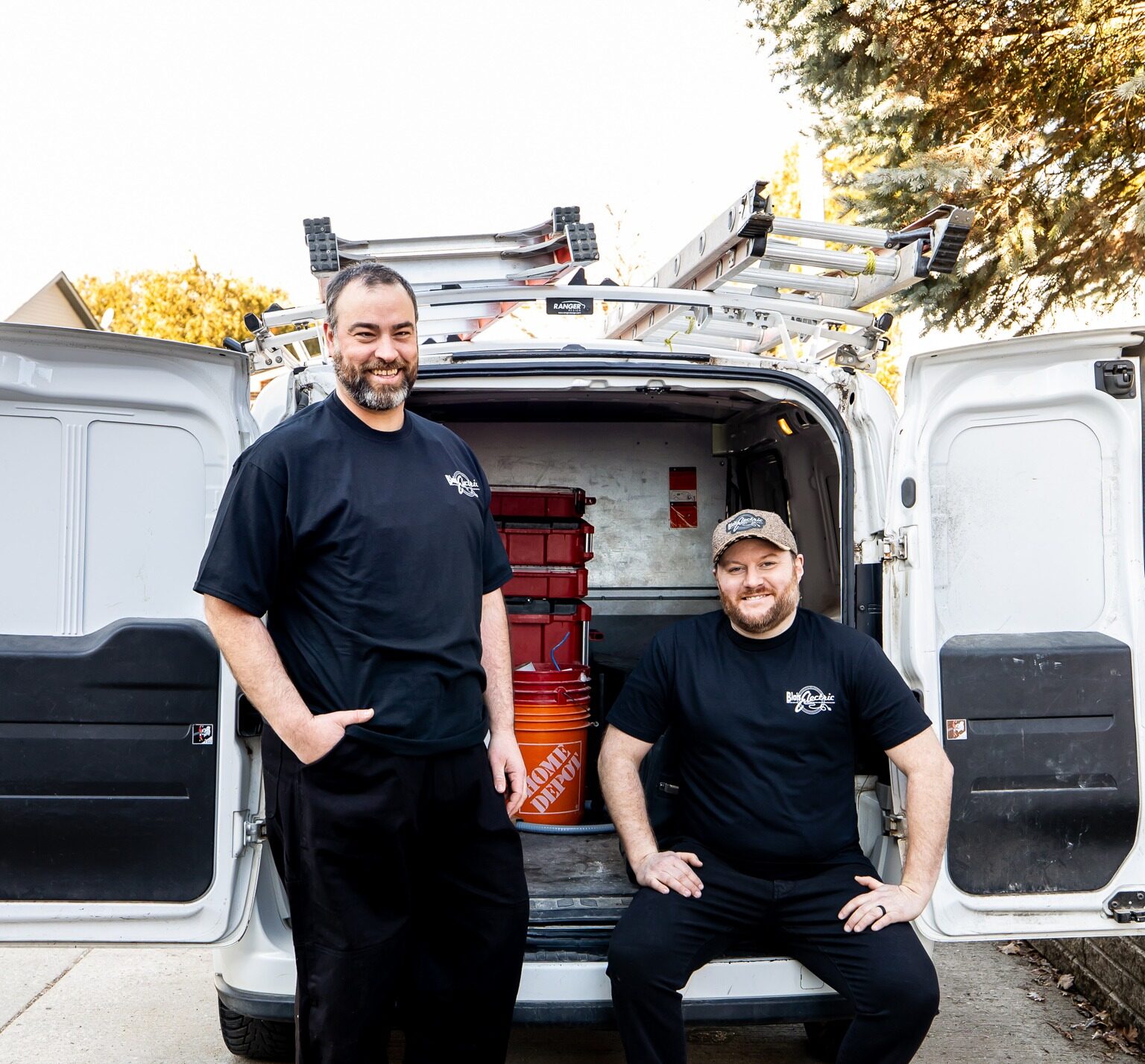 Two people are posing with a smile in front of an open utility van equipped with ladders and tools, suggesting they are professional service or trade workers.