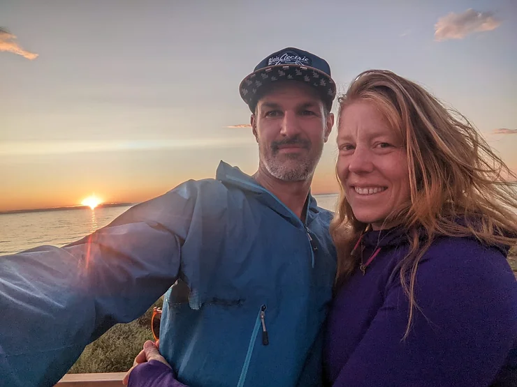 Two people are smiling for a selfie during sunset by the sea. Both are in casual attire, with a clear sky and calm waters behind them.