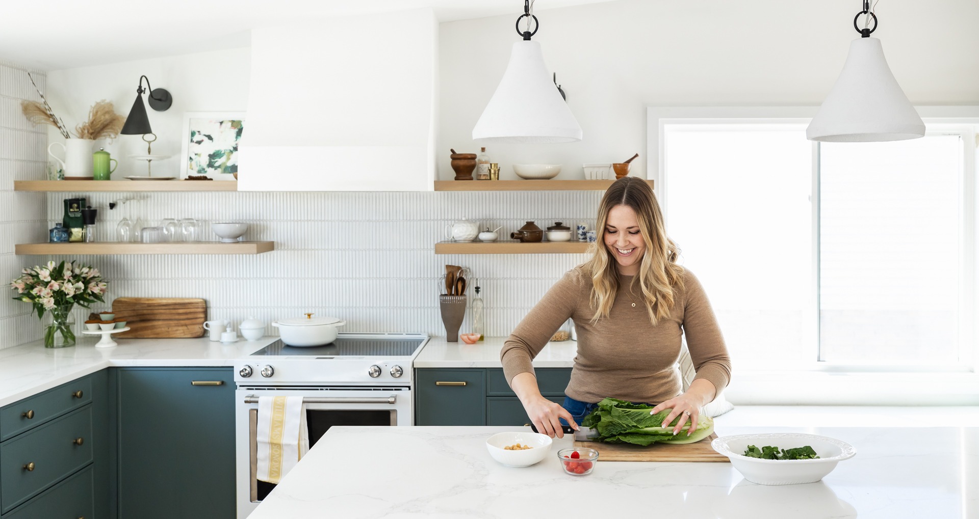 A person is preparing food in a modern kitchen with white subway tiles, open shelving, dark cabinets, marble countertops, and pendant lighting.