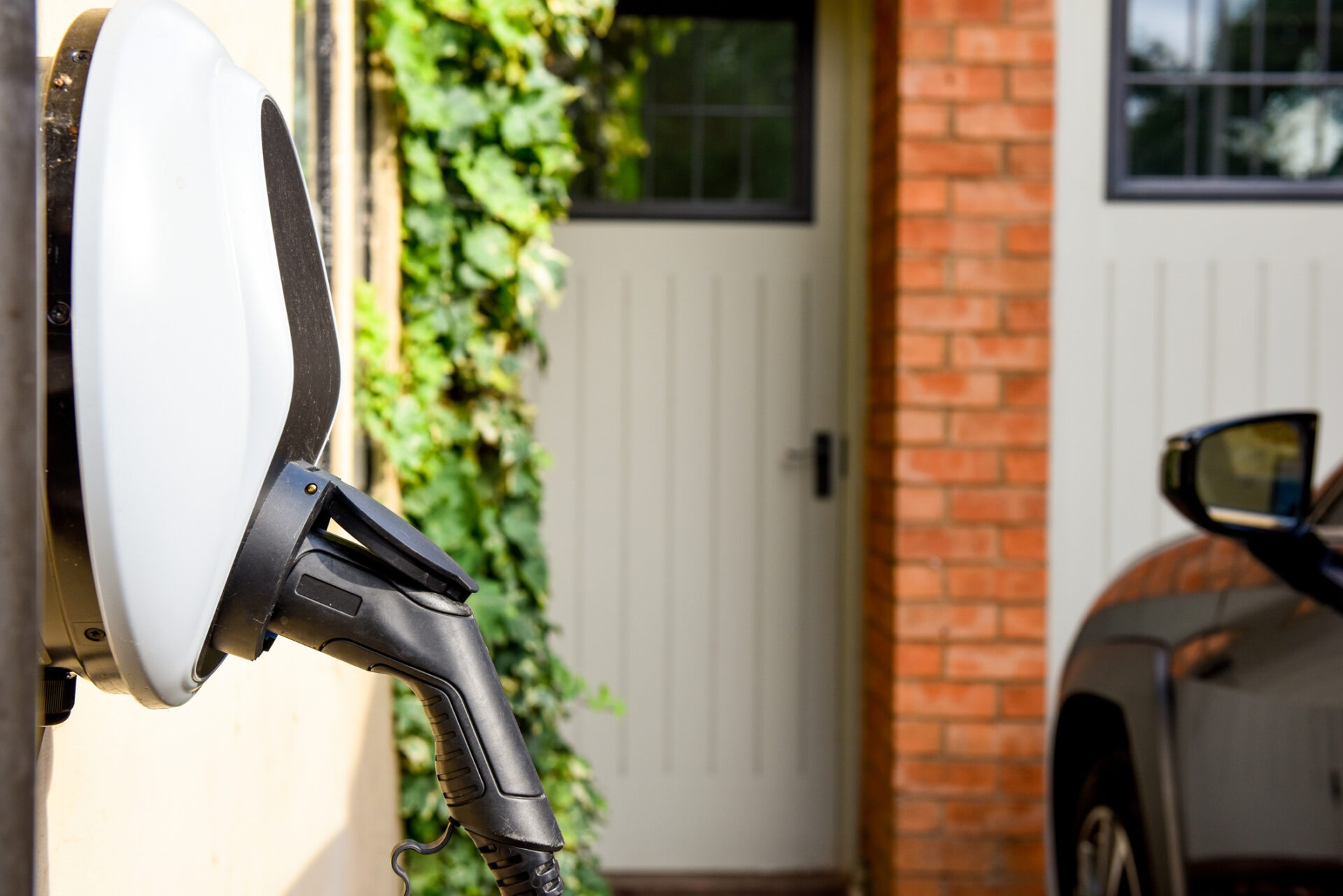 An electric vehicle charging station with a plug in the foreground, a partial view of a car, and a garage door surrounded by greenery and brick walls.