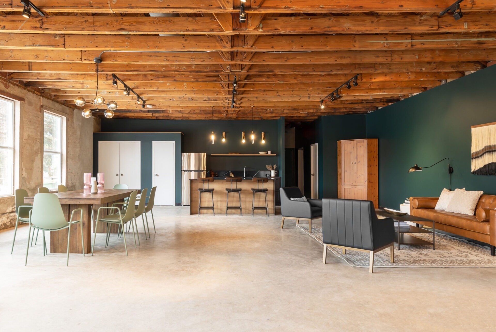 Modern industrial-style loft with exposed wooden ceiling beams, concrete floors, a dining area, a kitchen with bar stools, and a cozy sitting nook.