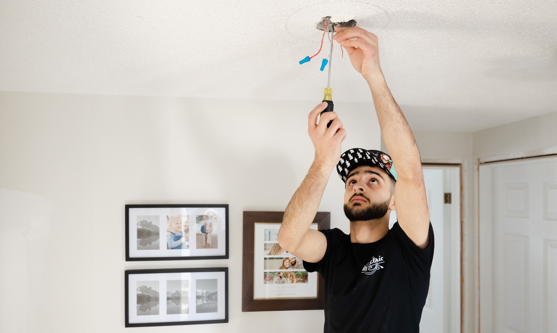A person is installing or fixing a ceiling light, holding a screwdriver and looking at exposed wires. There are framed pictures on the wall.