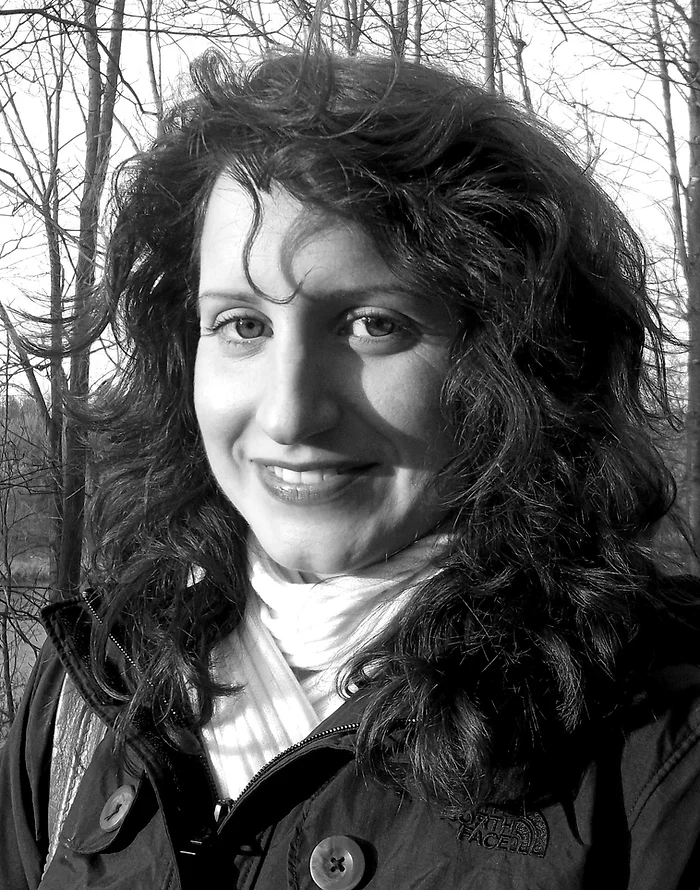 A black and white photo showing a smiling person with curly hair, wearing a jacket and scarf, with trees in the background.