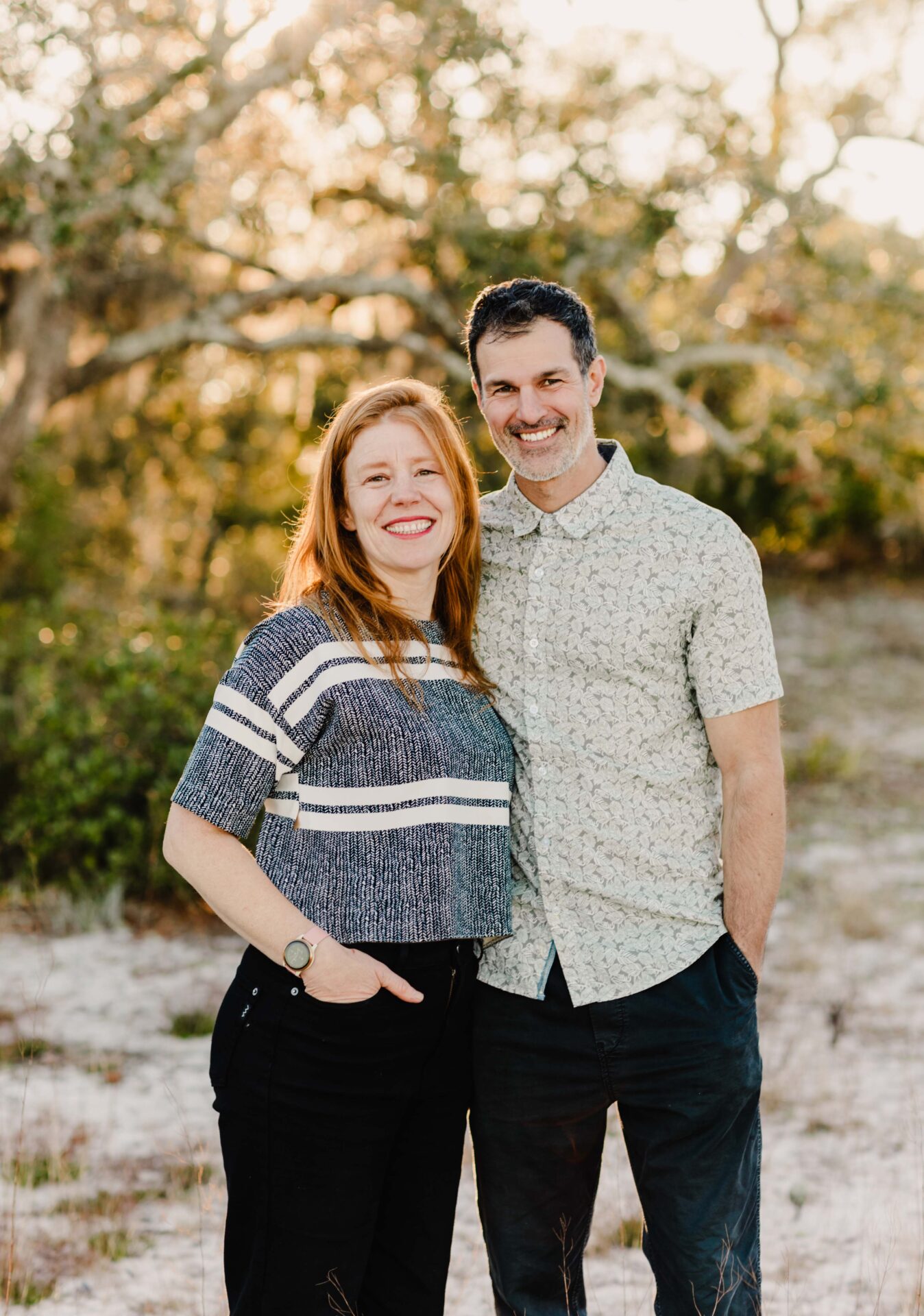 Two people stand embracing, smiling in front of a natural backdrop with trees and sunlight. They're dressed casually, conveying warmth and happiness.