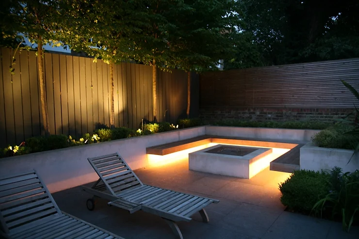An outdoor space at dusk features two loungers, ambient lighting around a central fire pit, wooden fences, a brick wall, and lush greenery.