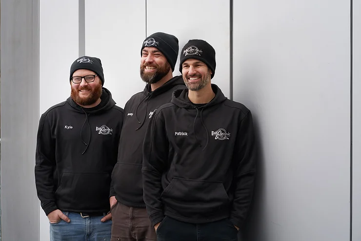 Three people are smiling and standing side by side against a grey background, wearing matching black hoodies and beanies with logos and names.