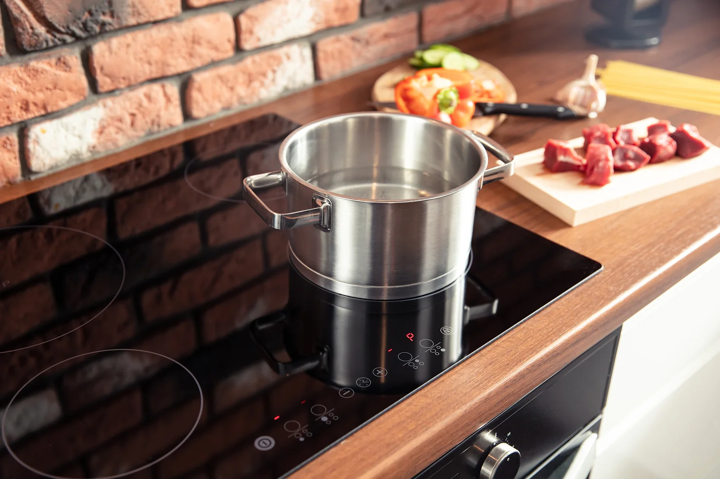 A stainless steel pot sits on an induction stove top in a kitchen with brick walls, beside ingredients like vegetables and meat.